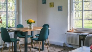 A dining area with a wooden table, four teal chairs, and a vase with yellow flowers. There are two windows with white frames, one on each side of the table. - Lovely Apartment in Straelen (Straelen)