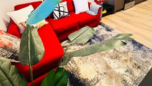 Un salon moderne avec un canapé rouge, une table basse ornée d’un motif en forme de feuille, et un coin cuisine doté de meubles en bois et d’une cuisinière.