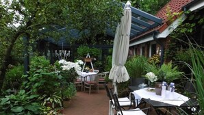 Un espace patio aménagé avec une table et des chaises, entouré de plantes en pot et agrémenté d’une pergola avec un parasol.