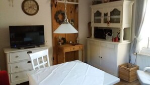 Une pièce comprenant un téléviseur posé sur un meuble, une table à manger recouverte d’une nappe blanche, un buffet en bois doté de portes en verre et un panier en osier sur le sol.
