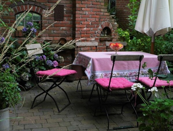En uteplats med ett bord och stolar, belagd med tegelstenar, och möblerna är täckta med rosa dukar. Uteplatsen är omgiven av växter och blommor.