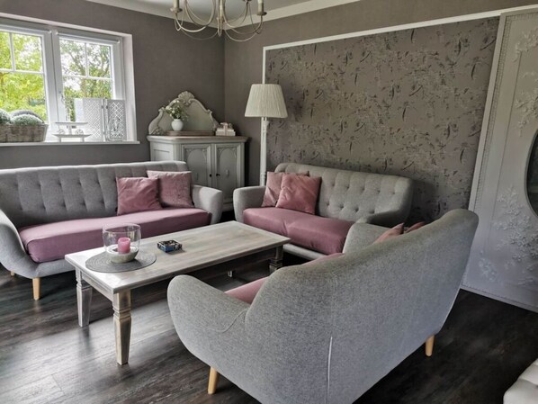 A living room with a grey sofa, a matching armchair, and a coffee table. There is a window with a view of greenery outside.
