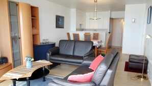 A living room with a sofa, a coffee table, and a dining area in the background. - Apartment in Nieuwpoort With Coastal Views (Nieuwpoort)
