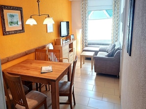 A room with a wooden dining table, chairs, a sofa, a television, and a window with patterned curtains. - Gemutliche Ferienwohnung auf der Insel Poel (Insel Poel)