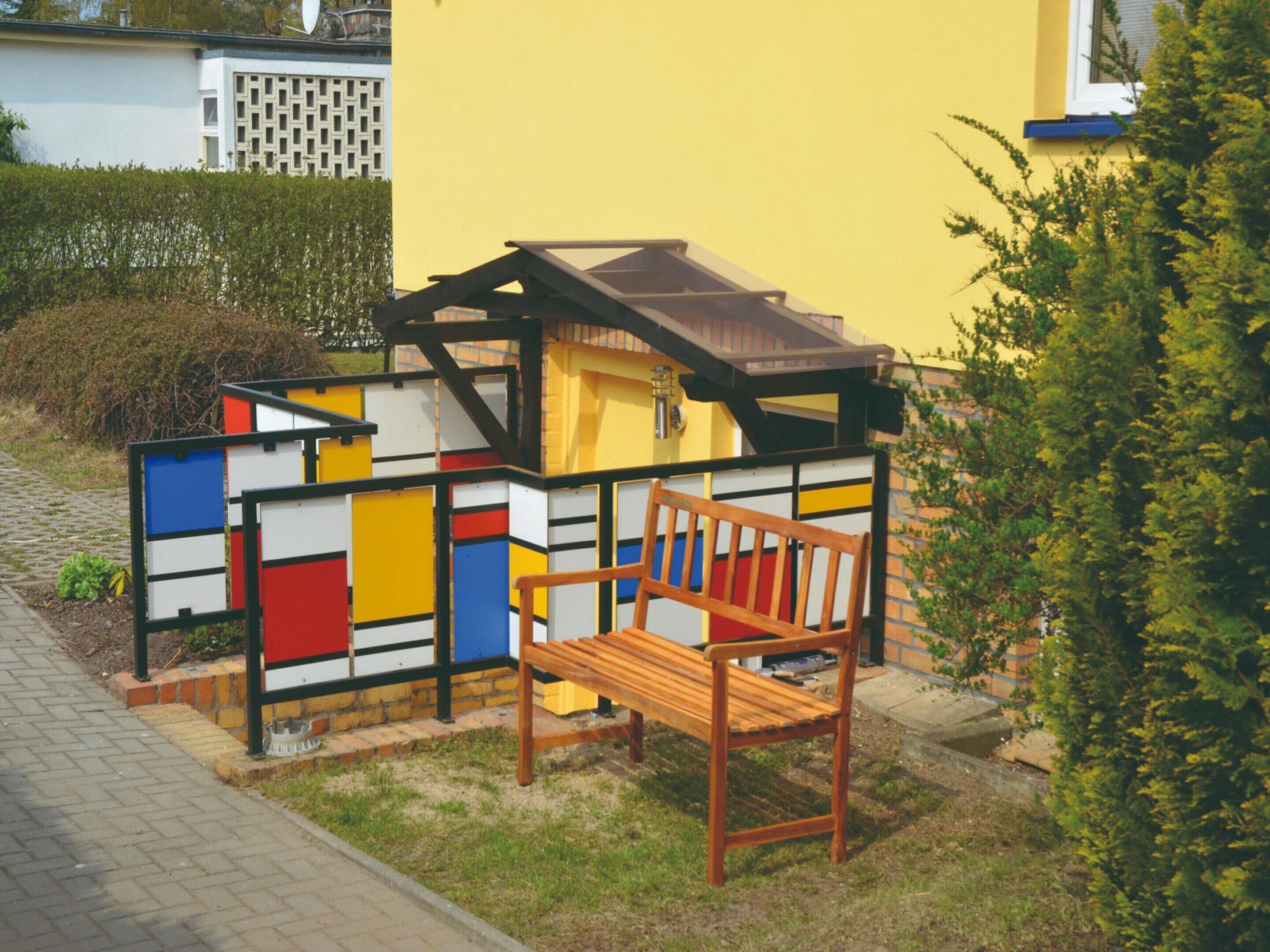 A colorful playhouse with a wooden bench in front, set against a yellow wall.