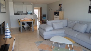 A modern living room with a grey sectional sofa, a small white table, and a colorful rug. There is a dining area with a wooden table and chairs, and a kitchen in the background. - Sea View Family Apartment (Nieuwpoort)