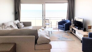 A modern living room with a sofa, armchairs, a TV, and a view of the sea through large windows. - Sea View Family Apartment (Nieuwpoort)