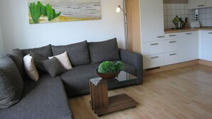 A modern living room with a grey sofa, a glass coffee table with a plant, and a kitchen in the background. - - 2 min zum Strand (Borkum)