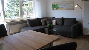 A modern living room with a sofa, a coffee table, and a dining table. There is a window with a view of trees and a painting on the wall. - - 2 min zum Strand (Borkum)