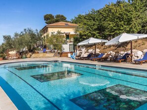 A clear swimming pool with lounge chairs and umbrellas, surrounded by trees and a building with a tiled roof. - Belvilla by OYO Villa Sandra (Mondavio)