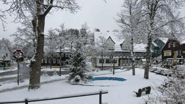 Um parque nevado com bancos, um pequeno lago e edifícios ao fundo.