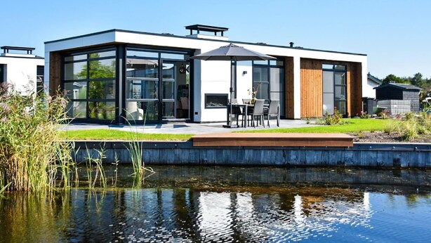 A modern house with large glass windows, a patio with outdoor furniture, and a pond in the foreground. - Loosdrecht Water Villa (Loosdrecht)