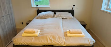 A double bed with two pillows and two towels, a window with a view of houses, a wooden floor, and a dark headboard.