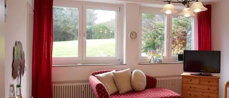A cozy living room with a red sofa, a wooden table, a TV, and large windows with red curtains.