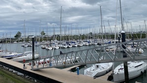 Ein Yachthafen mit zahlreichen Segelbooten, einer Fußgängerbrücke und einer grünen Fläche im Hintergrund.