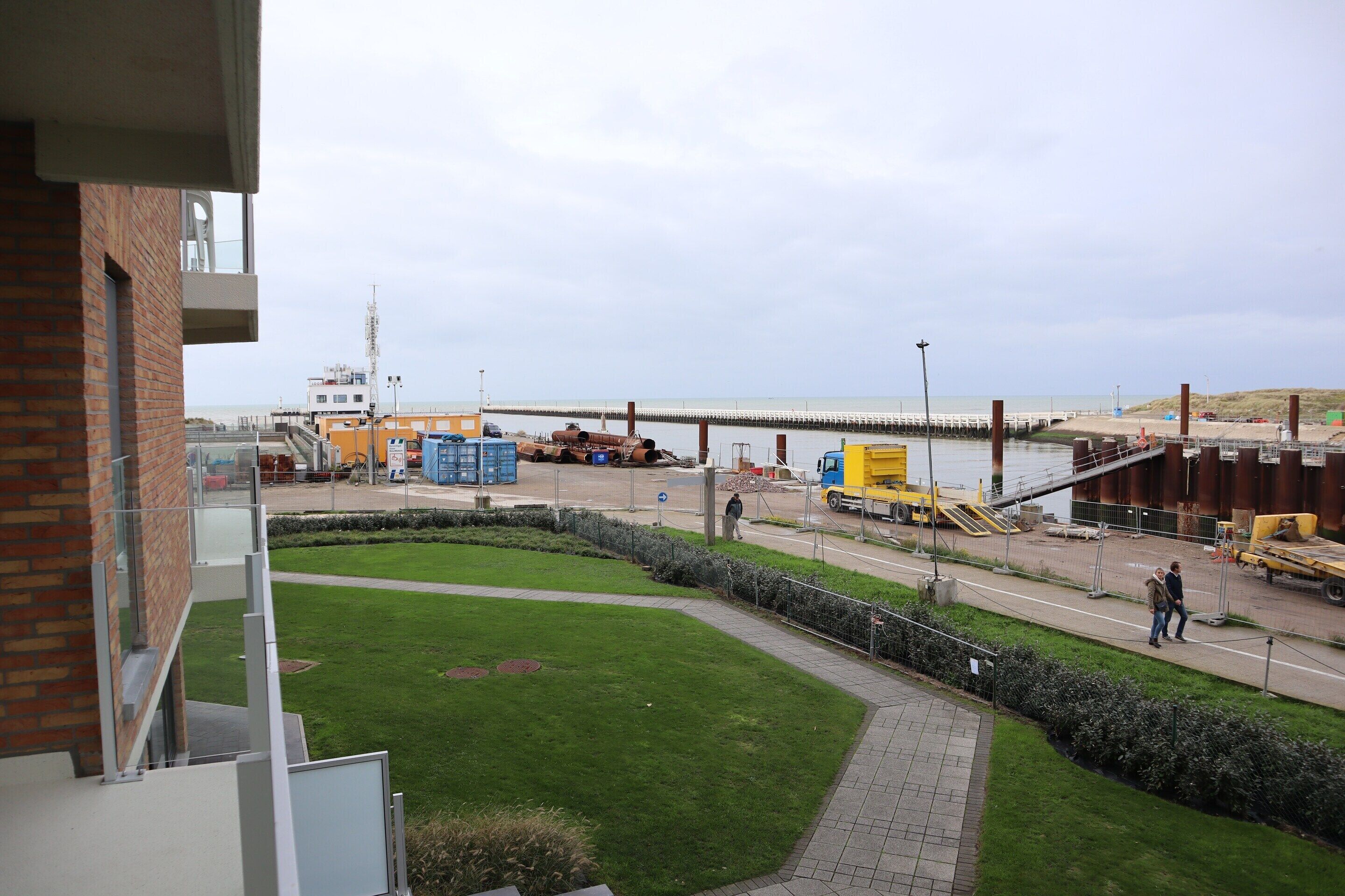 A view from a balcony overlooking a construction site with a bridge, a grassy area, and a paved walkway.