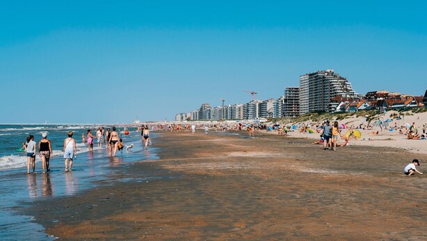 Ein Strand mit Menschen, die am Ufer entlanggehen, einem klaren Himmel und Gebäuden im Hintergrund.