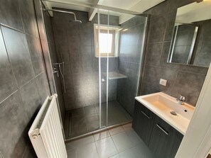 Une salle de bain moderne avec une douche à l’italienne en verre, un lavabo blanc et des murs recouverts de carreaux foncés.