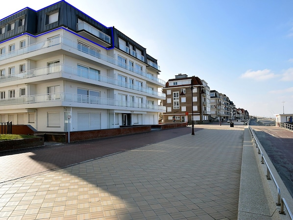 Un edificio moderno de varios pisos con balcones, un sendero pavimentado y un cielo despejado.