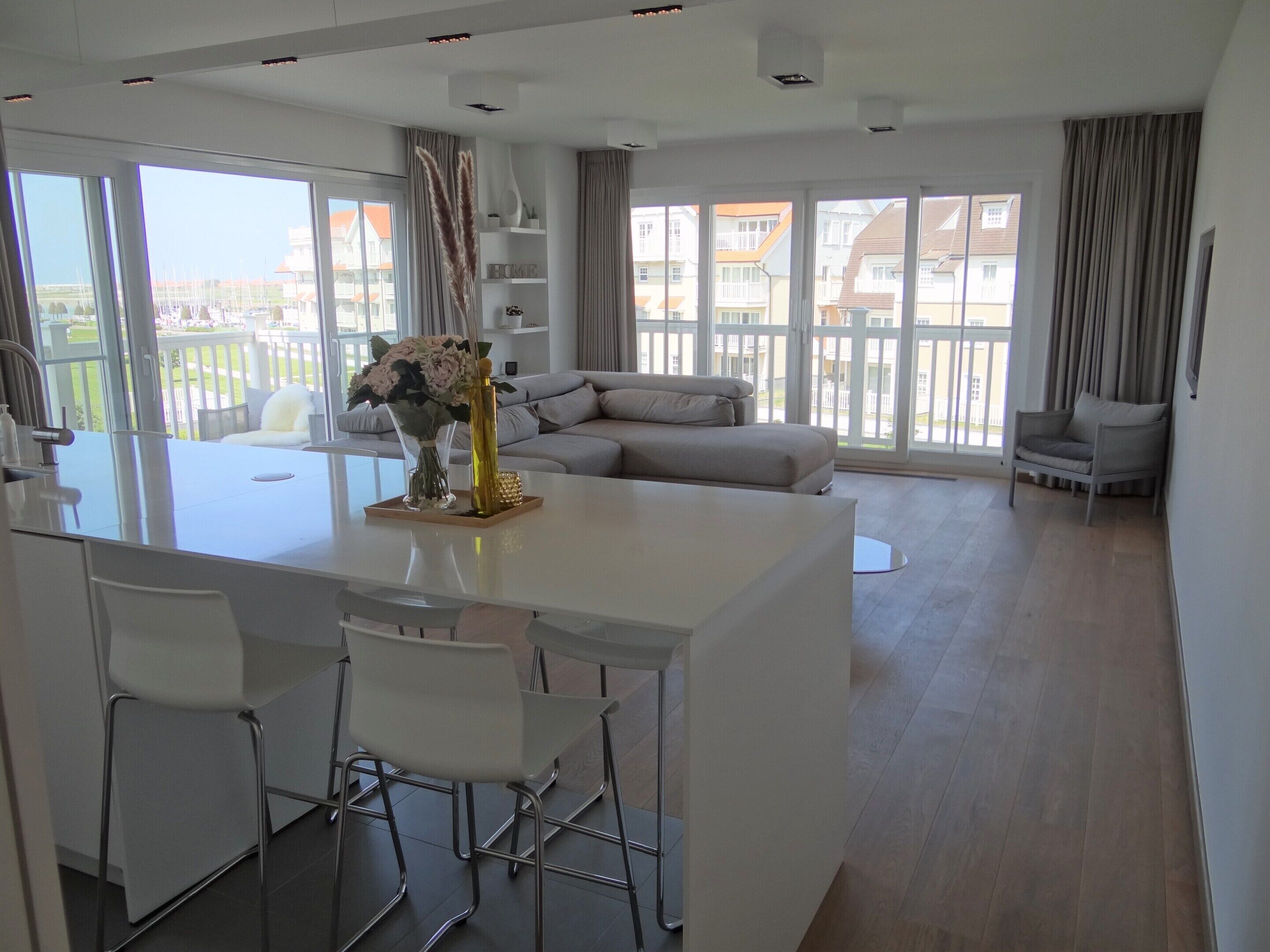 A modern living area with a central island, white furniture, and large windows offering a view of the city.