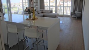 A modern living area with a central island, white furniture, and large windows offering a view of the city. - Apartment in Nieuwpoort Near the Beach (Nieuwpoort)