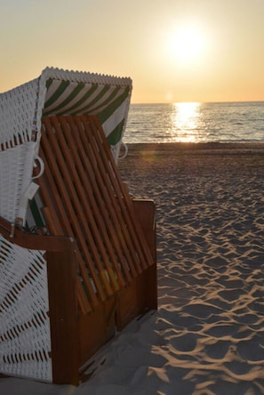 A beach chair with a striped cover and a wooden frame, positioned on a sandy beach with the sun setting in the background. - Our-island-scharbeutz (Scharbeutz)
