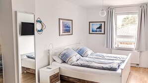 A bedroom with a bed, bedside table, and a mirror. There are framed pictures on the wall and a window with curtains. - Our-island-scharbeutz (Scharbeutz)