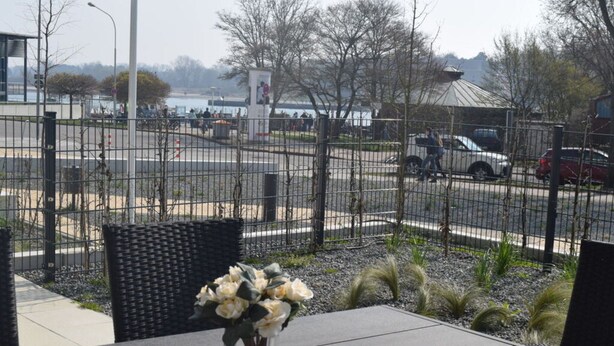 A table with a vase of flowers, a wicker chair, and a view of a parking lot with cars and a building in the background. - High End Travemunde Comfortable Vacation Home (Lübeck)