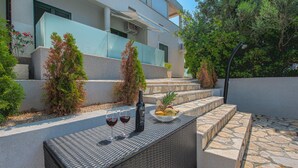 A modern outdoor seating area with a stone-tiled floor, a wicker table, and a bowl of fruit and a bottle of wine. - Cosy Apartment in Pag (Pag)