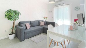 A modern living room with a grey sofa, a dining table with chairs, a potted plant, and a large window. - Cosy Apartment in Pag (Pag)