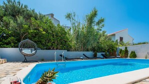A swimming pool with lounge chairs and a hanging chair, surrounded by greenery and a stone patio. - Cosy Apartment in Pag (Pag)