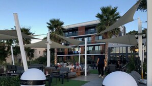 An outdoor lounge area with seating and a bar, surrounded by palm trees and modern architecture. - Apartment Oleander (Novalja)