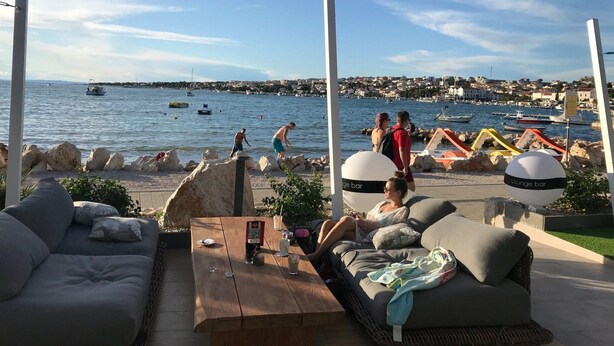 An outdoor seating area with a view of the sea, a lounge bar, and a rocky shoreline. - Apartment Oleander (Novalja)