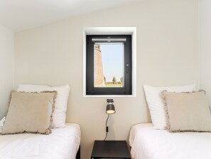 Two single beds with beige pillows and white bedding, a bedside table with a lamp, and a window with a view of a building and trees.