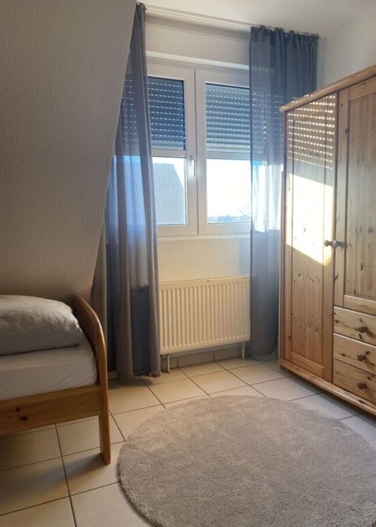 A bedroom with a bed, a radiator, a wardrobe, and a window with curtains.