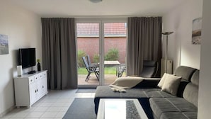 Un salon moderne comprenant un canapé, une table basse, un téléviseur posé sur un meuble et une porte-fenêtre coulissante donnant sur une terrasse extérieure avec une table et des chaises.