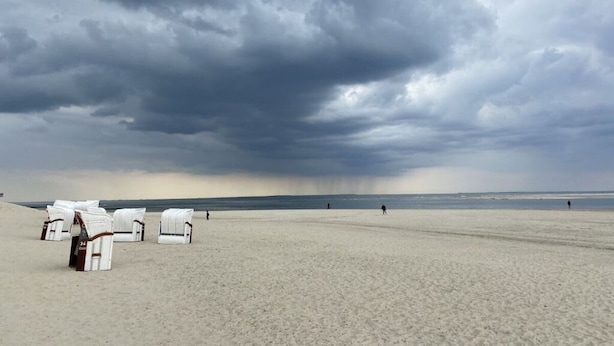 Une plage avec des chaises longues et des parasols blancs, un rivage sablonneux et un ciel orageux.