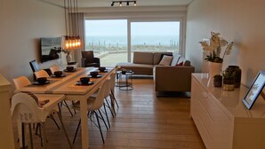 A modern living room with a dining area, large windows, and a view of the beach. - Apartment in Nieuwpoort With sea View (Nieuwpoort)