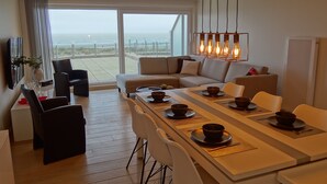 A modern living room with a large dining table set for two, a sofa, and a view of the beach. - Apartment in Nieuwpoort With sea View (Nieuwpoort)