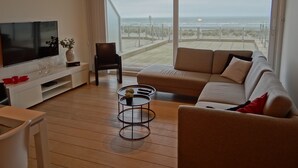 A modern living room with a large TV, a sofa, a chair, and a small table. There is a large window with a view of the beach. - Apartment in Nieuwpoort With sea View (Nieuwpoort)