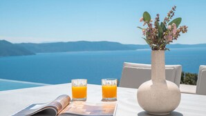 Ett bord med en vas med blommor, två glas apelsinjuice och en tidning på en terrass med utsikt över en vattenyta.