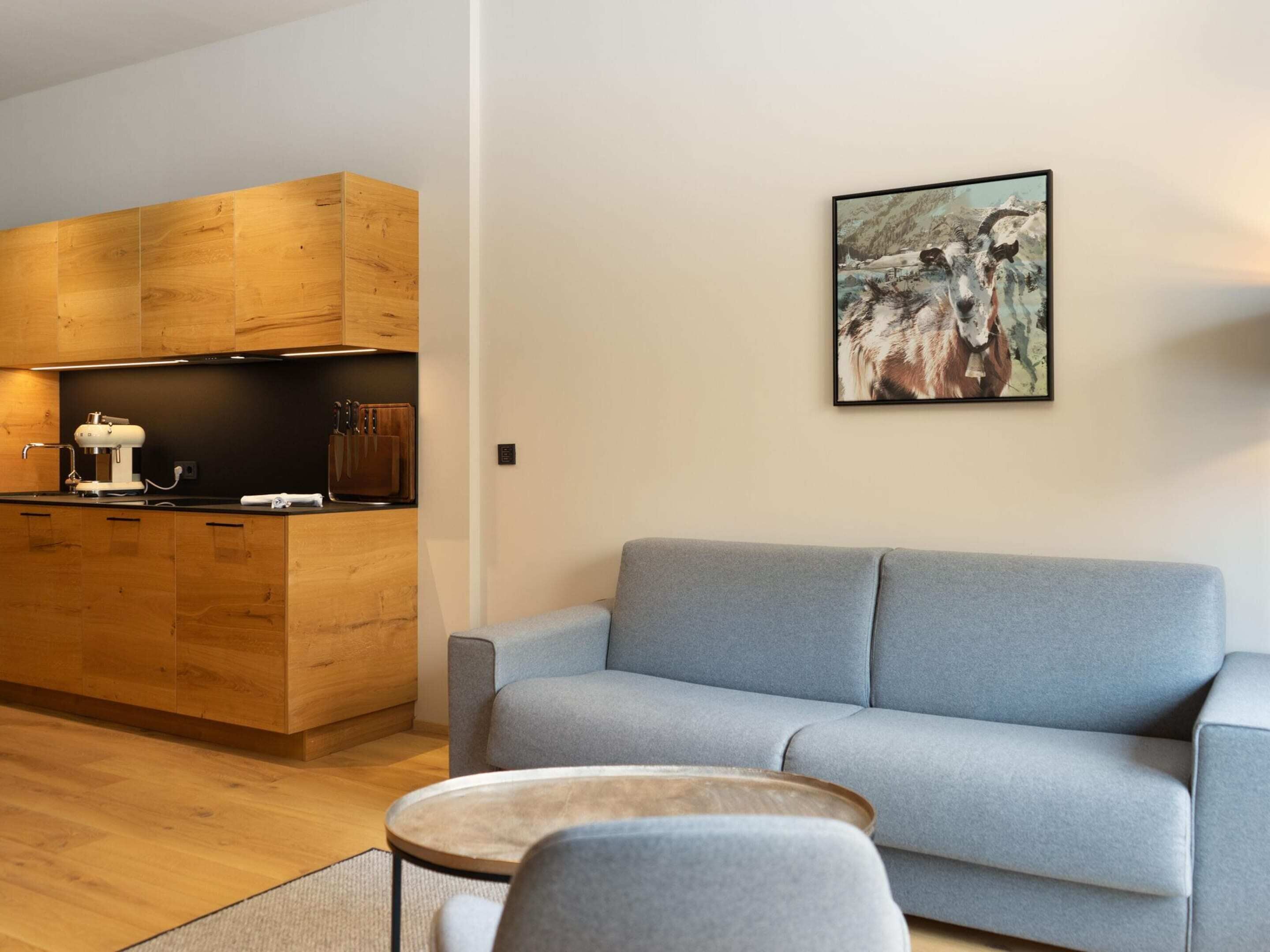 A modern kitchen with wooden cabinets and a built-in oven, a gray sofa, and a small round table.