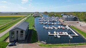 A marina with boats docked, a building with a sloped roof, and a grassy area with a pathway. - Apartment Kamperland With Harbour View (Kamperland)