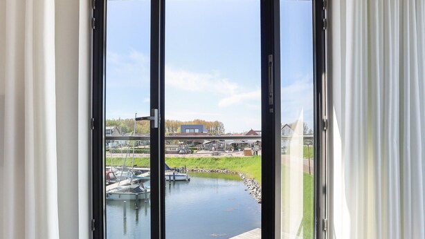 A view through a glass door to a water body with boats, a dock, and a building in the background. - Apartment Kamperland With Harbour View (Kamperland)