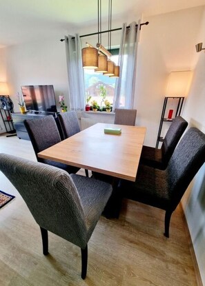 A modern living room with a wooden dining table, grey upholstered chairs, a flat-screen TV, and a window with curtains. - Uitzicht op Staufen (Piding)