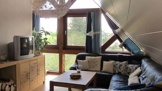 Een gezellige woonkamer met een zwarte leren bank, een houten salontafel en een televisie op een houten kast. Grote ramen laten natuurlijk licht en uitzicht op groen binnen.