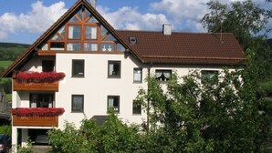 Een tweelaags huis met een schuin dak, een balkon met rode bloemen, en een auto op de oprit.