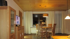 Een gezellige woonkamer met een houten kast, een televisie, een eettafel met stoelen en een raam met gordijnen.