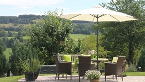 Een terras buiten met een tafel en stoelen, omgeven door groen en uitzicht op een glooiend landschap.