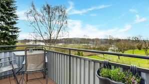A balcony with a table and chairs, a metal railing, and a view of a green landscape with trees and a body of water. - Apartment in Holstein With Balcony (Ploen)